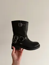 Bota de caña media en eco cuero negro, con diseño estilo biker que incluye correas con hebillas metálicas y herrajes plateados en el tobillo. Cuenta con suela plana y punta redondeada.