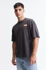 Remera de manga corta color gris oscuro, con corte holgado, hombros caídos y escote redondo. Presenta un logo estampado en el pecho con la inscripción "GALLERY DEPT." en color naranja.