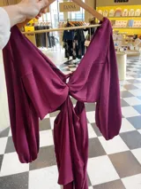 Vestido largo color bordó con escote en V profundo, detalle de nudo central y aberturas laterales en la cintura. Presenta mangas largas abullonadas con puños elásticos y falda con tajo frontal pronunciado.