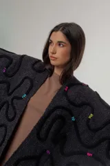 Ruana de tejido de punto grueso en color gris oscuro, con diseño de líneas curvas en relieve de color negro y detalles de costuras decorativas en colores vibrantes. Presenta terminaciones con flecos en el ruedo y una etiqueta de cuero sintético en la parte inferior.