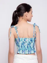 Top corto con escote recto y tirantes finos ajustables mediante nudo en los hombros. Presenta un estampado floral sobre fondo oscuro, con flores rosadas y hojas turquesas.