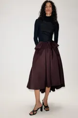 Falda midi asimétrica de color bordó, confeccionada en tejido satinado con volumen. Presenta un diseño con volados en la cintura y cierre de cremallera oculta en la espalda.