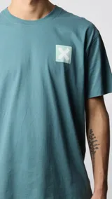 Camiseta de manga corta color verde azulado, corte holgado, con un pequeño estampado cuadrado con una 'X' en el pecho.