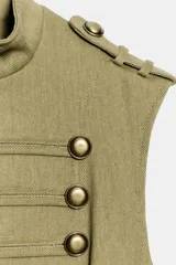 Chaleco entallado color verde militar de cuello subido y manga sisa, con trabillas en los hombros. Cuenta con detalle de hileras horizontales de cordoncillo y botones metálicos dorados en el delantero. Forro interior y cierre frontal con corchetes ocultos.