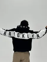 Canguro con capucha de corte boxy, color negro con una franja horizontal blanca que recorre la espalda y las mangas, presentando la palabra 'BROOKLYN' estampada en negro sobre la franja. La capucha también incluye detalles estampados.