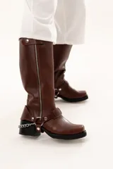 Bota de caña alta estilo biker confeccionada en cuero color chocolate. Presenta un cierre metálico lateral que recorre toda la caña, taco cuadrado y plataforma. Incluye un sistema de estribo y cadena metálica desmontable en la zona del tobillo, permitiendo diversas formas de uso.