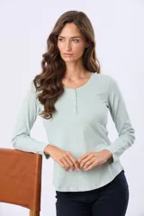 Remera de manga larga en color verde menta, con cuello redondo y cierre frontal parcial de botones a tono. Presenta un diseño con ruedo curvo y un pequeño aplique metálico en la parte inferior.