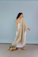Ruana tejida a mano en lana merino color beige, con textura de punto y detalle de flecos en el ruedo. Presenta sutiles destellos brillantes integrados en el tejido.