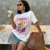 Remera blanca de corte holgado con estampa frontal colorida que incluye rayas verticales rosadas y un diseño gráfico de un trago con rodaja de naranja. Confeccionada en mezcla de algodón y poliéster.