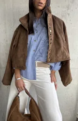 Camisa clásica de manga larga, a rayas verticales azules y blancas, con cuello camisero y botones frontales. Presenta un bordado lineal de un rostro en el frente.