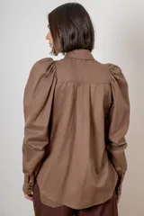 Camisa de algodón color marrón con mangas largas abullonadas, cuello clásico y cierre frontal oculto. Presenta botones decorativos con strass en los puños y en el cuello.