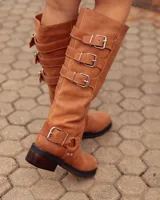 Bota de caña alta confeccionada en cuero color marrón tostado. Presenta un diseño con tres hebillas decorativas en la parte superior de la caña y una correa con hebilla y argolla metálica en el tobillo. Cuenta con suela plana y taco bajo de madera oscura.