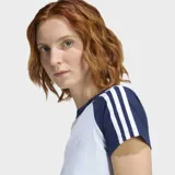 Remera Adidas de ajuste ceñido, color celeste claro con mangas cortas en azul marino y las icónicas tres rayas blancas en los hombros. Presenta cuello redondo acanalado y el logo del trébol bordado en el pecho.