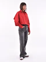 Camisa cropped color rojo con cuello clásico, botones negros en el frente y mangas largas con puños abotonados. Presenta un detalle de abertura en la parte superior de las mangas.