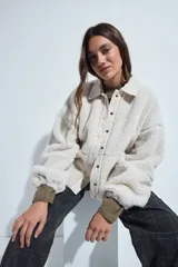 Campera de corderito color beige, con cuello camisero y cierre frontal de botones a presión. Presenta una cartera central lisa en contraste y mangas largas con puños elásticos.
