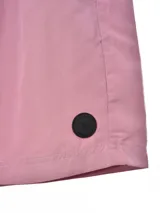 Short de baño color rosado, con cintura elástica y cordón de ajuste blanco. Presenta bolsillos laterales y un pequeño parche circular negro en la parte inferior derecha.