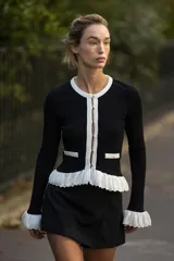 Chaqueta de punto acanalado en color negro con detalles en blanco en cuello, tapeta de botones, bolsillos y volados en el ruedo y puños. Presenta cierre frontal con botones dorados.