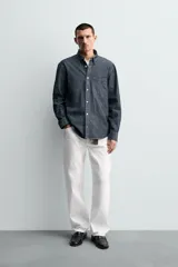 Camisa de corte relajado confeccionada en tejido de algodón denim, con cuello solapa, manga larga con puños abotonados y bolsillo plastrón en el pecho. Presenta cierre frontal de botones y bajo con aberturas laterales.