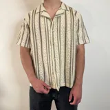 Camisa de manga corta con cuello cubano, confeccionada en tejido de crochet con diseño de rayas verticales en tonos beige y marrón. Presenta un corte boxy y cierre frontal con botones.