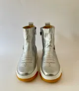 Botas cortas de cuero plateado, con diseño híbrido que combina la estética texana con una suela chunky blanca y detalle de arte lateral creado por Carolina De Cunto.