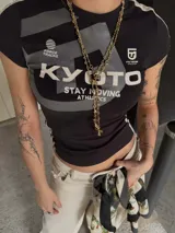 Top de manga larga con diseño superpuesto, que combina una remera de manga corta negra con estampado gráfico frontal y mangas largas blancas debajo. Presenta un ajuste al cuerpo y estilo deportivo.