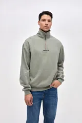 Buzo de felpa con friza, color verde oliva, con cuello alto y cierre frontal parcial. Presenta mangas largas con puños elásticos, ruedo acanalado y bordado de la marca en el pecho.