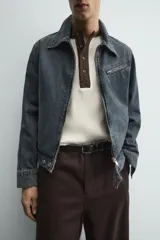 Cazadora cropped fit de denim de algodón, color negro lavado. Presenta cuello solapa, manga larga con puños abotonados, cierre frontal de cremallera y bolsillo superior con cierre de cremallera en el pecho.