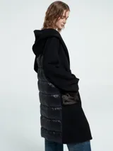Abrigo largo de paño negro con capucha, cierre cruzado con lazo ajustable en la cintura y bolsillos delanteros con textura de pelo sintético. Presenta un diseño con espalda en nylon capitoneado.