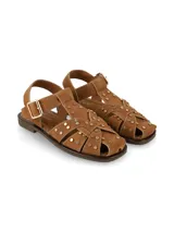 Sandalia plana tipo guillermina y cangrejera, confeccionada en cuero gamuza color camel, con tiras cruzadas decoradas con tachas metálicas doradas. Cuenta con tira ajustable en el talón con hebilla metálica y suela de caucho.