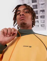 Campera deportiva tipo track jacket, color amarillo mostaza con detalles en verde oscuro. Presenta cuello y puños acanalados, cierre frontal y tres franjas contrastantes en las mangas. Pequeño logo bordado en el pecho.