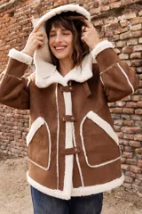 Campera estilo gamulán color tostado con interior y bordes de corderito sintético color crema. Presenta capucha amplia, cierre frontal con trabas y broches metálicos, y bolsillos delanteros grandes. Posee un calce relajado y diseño de textura suave.