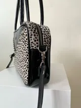 Cartera rígida negra con detalles en rojo y tachas plateadas. Tiene herrajes y cierre metálico plateado marca YKK, asa fina para llevar al hombro o en la mano y correa larga extra regulable. El interior es forrado, tiene un bolsillo con cierre enmarcado en cuero, y un segundo bolsillo plaqué.