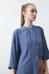 Camisa de color celeste claro con silueta relajada y mangas largas. Presenta un cuello redondo con pequeña abertura frontal y botones, además de un delicado trabajo de tablas en los hombros que aporta textura al diseño.