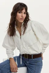 Blusa de algodón liviano color blanco, con detalles de puntilla en el frente, cuello mao con escote en V, cordón colgante y mangas largas abullonadas con puños ajustados.