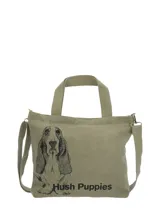 Cartera tipo tote bag de canvas en color verde oliva, con estampado frontal de un perro basset hound y logo de la marca. Cuenta con doble asa de mano, correa ajustable extraíble, cierre principal de cremallera y bolsillos internos y traseros con cierre.