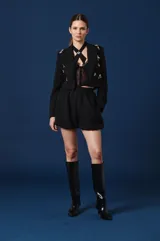 Saco tipo spencer negro de corte corto y estructurado, con solapas clásicas y detalles de pines decorativos distribuidos en la parte frontal.