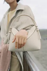 Bolso de mano formato mini saca, color crema, con diseño estructurado de líneas geométricas y costuras visibles. Incluye asa de mano fija y correa bandolera extraíble.