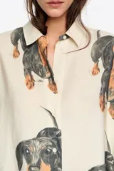 Camisa de corte recto con cuello clásico, cierre frontal de botones y manga larga con puños abotonados. Presenta un estampado integral de perros salchicha sobre fondo color crema.