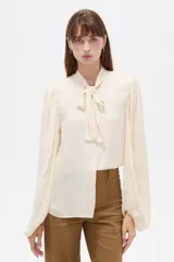 Camisa de poliéster color marfil, de diseño holgado, con cuello alto lazada y mangas largas abullonadas.