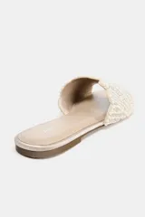 Sandalia plana tipo pala de color beige, con tira ancha decorada con perlas y abalorios brillantes dispuestos en patrón radial, punta redonda y suela plana.