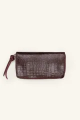 Billetera rectangular de cuero con textura de reptil, cierre perimetral y correa de muñeca lateral. Presenta un pequeño aplique metálico circular en el centro.