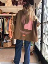 Campera de corderito marrón con capucha, diseño de apliques geométricos abstractos en tonos rosados y bordó en la parte posterior, y etiqueta frontal de la marca.