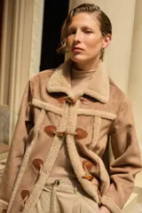 Abrigo de estilo duffle coat en simil gamuza color tostado, con interior y bordes de piel sintética al tono. Presenta cierre frontal mediante alamares de cuero sintético y madera, cuello solapa y mangas largas.