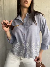 Camisa celeste a rayas verticales blancas, con cuello clásico, cierre frontal con botones, mangas largas con puños abotonados y detalle de bordado floral en el ruedo.