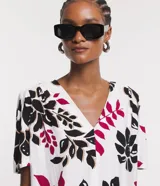 Blusa de viscosa con escote en V y mangas cortas abullonadas. Presenta un estampado de follaje y flores en tonos negro, fucsia y beige sobre fondo blanco.