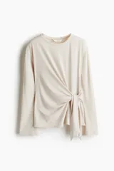 Top de manga larga de punto suave color beige, con diseño de hombros caídos, escote redondo y detalle de tiras para anudar en el lateral de la cintura.