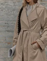 Trench coat de corte midi en color beige, con cuello de solapa amplio, cierre frontal con cinturón ajustable a la cintura, bolsillos laterales y detalle de botones en los puños.