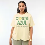 Remera de corte oversize en color amarillo claro, con cuello redondo y mangas cortas. Presenta un estampado gráfico frontal con la frase 'COSTA AZUL' en letras verdes y un diseño de sol, acompañado por el texto 'Palma de Majorque' en tipografía cursiva.
