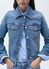 Chaqueta de jean estilo crop, confeccionada en algodón con un toque de elastano, de diseño recto y color azul. Presenta cuello camisero, cierre frontal con botones metálicos, mangas largas con puños abotonados y dos bolsillos de parche con solapa en el pecho.