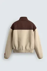 Cazadora de corte relajado confeccionada en tejido de borreguillo color beige combinado con tejido técnico marrón en la parte superior. Presenta cuello subido, cierre frontal con cremallera, bolsillos laterales de vivo y puños y bajo elásticos. Incluye parches y bordados decorativos en el pecho.